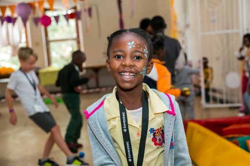 A smiling girl with a painted face stands in the foreground of children in a classroom setting