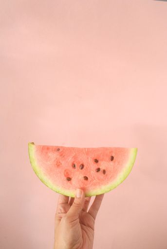A hand of a person holding  a slice of watermelon in a pink background
