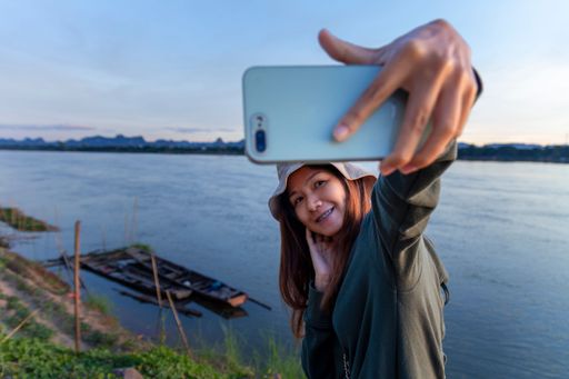 A young woman taking a photograph with a modern white smartphone of a panoramic view of a tranquil river surrounded by lush green trees