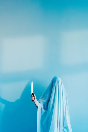 A vertical shot of a person covered with blue fabric holding a sharp knife against blue wall