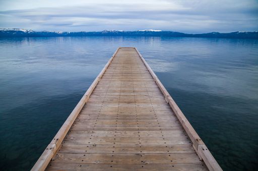 A wooden dock over the calm blue waters of Lake Tahoe with snow capped mountains in the distance