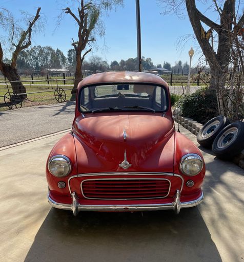A photo of a front view of a really rare small British Minor 1000 Classic Car parked outside on driveway with a blue sky & trees in the background
