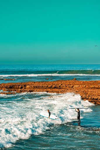 A vertical shot of two surfers walking towards the waves of the beach, carrying their surfboards