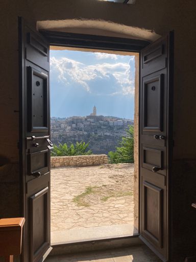 A vertical shot of an open wooden door with a view of a hill with buildings on a sunny day