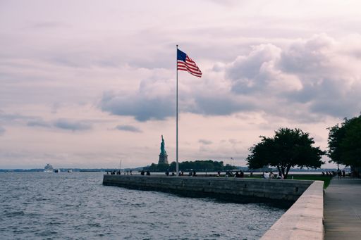 A selective focus shot of a U.S. flag with the Statue of Liberty in the background and ships sailing in Upper Bay, on a beautiful cloudy day