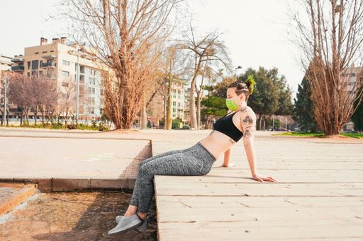 A sexy Spanish woman in a face mask with tattoos resting after exercise in the park - new normal concept