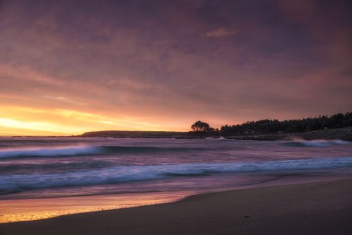 A mesmerizing view of the wavy ocean hitting the sandy beach gleaming under the sunset in the clouds