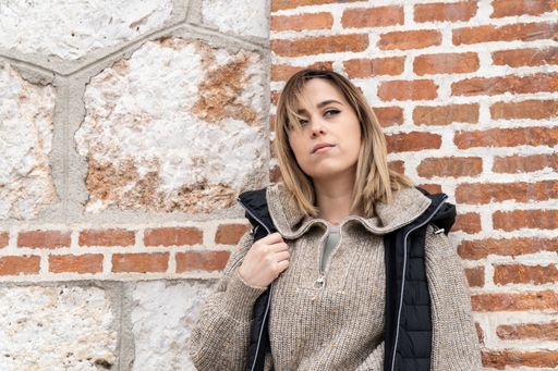 Horizontal shot of a straight-haired blonde woman in a brown sweater and black vest in front of a stone and brick wall.