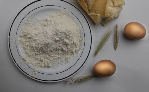 An overhead shot of flour in a plate and eggs and bread next to it on a white a table