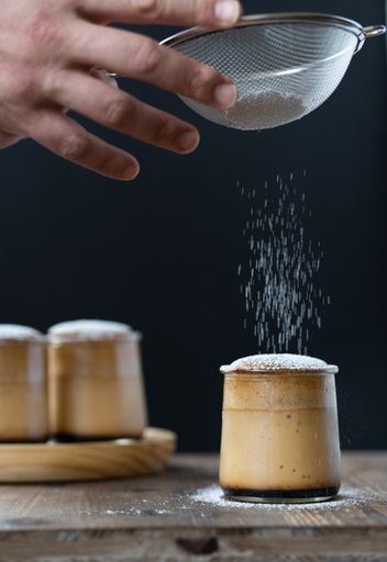 A vertical shot of a baker sprinkling icing sugar over Japanese style flan made with eggs and honey, and topped with sponge cake