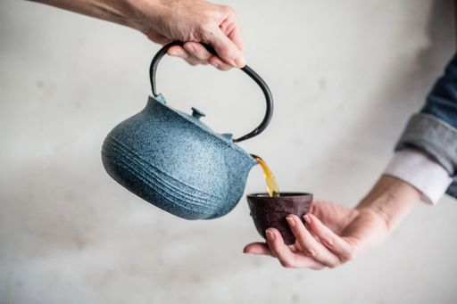 A hand holding a blue teapot pouring out tea into a cup in another person's hand