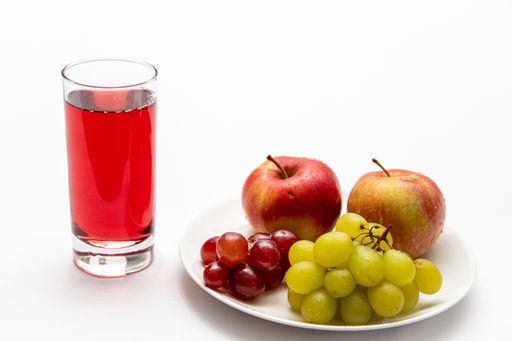 A closeup of a glass of red fruit juice and a plate with fresh apples and grapes on white background