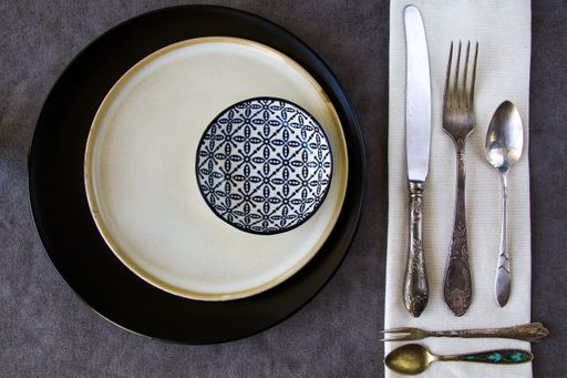 A top view of an empty ceramic bowl on plates with a fork, a knife, and spoons on the table