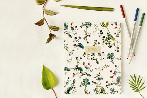 A closeup top view of a notebook with floral ornaments cover surrounded by colorful pencils and plant leaves