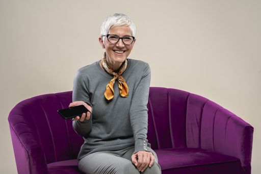 A cheerful middle-aged Caucasian female sitting on a purple couch using a phone and smiling against a white background