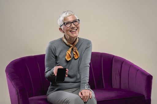 A cheerful middle-aged Caucasian female sitting on a purple couch using a phone and smiling against a white background