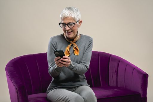 A cheerful middle-aged Caucasian female sitting on a purple couch texting on the phone and smiling against a white background