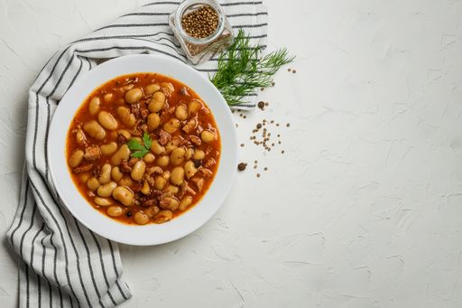 A white ceramic bowl filled with a soup of beans and meat on a light table with greens and peppercorns.