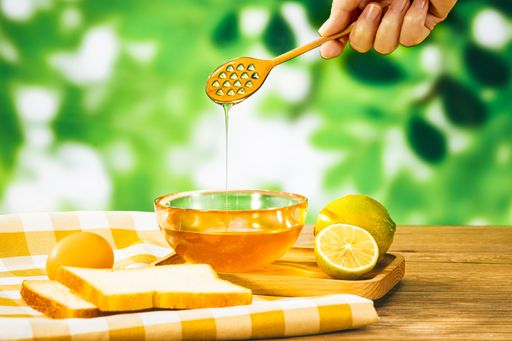 A closeup of a hand holding a wooden dipper with honey pouring into a bowl next to rustic products
