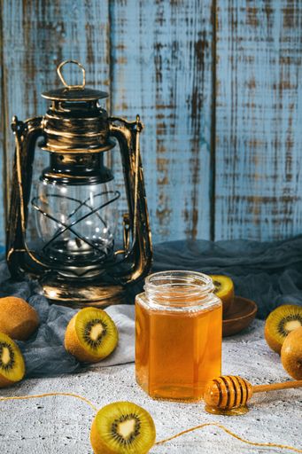 A vertical shot of a wooden dipper with honey surrounded by kiwis next to a lantern