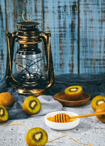 A vertical shot of a wooden dipper with honey surrounded by kiwis next to a lantern