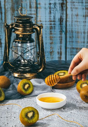 A vertical shot of a hand holding a wooden dipper with honey surrounded by kiwis next to a lantern