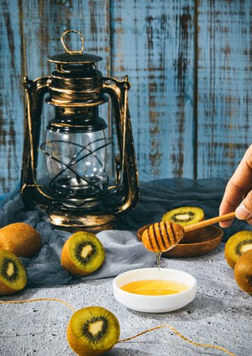 A vertical shot of a hand holding a wooden dipper with honey surrounded by kiwis next to a lantern