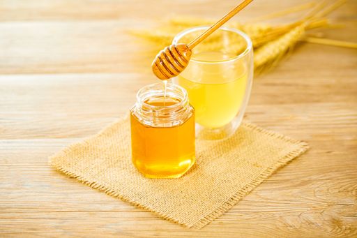 A closeup of golden honey pouring from a wooden dipper into a jar next to a juice glass