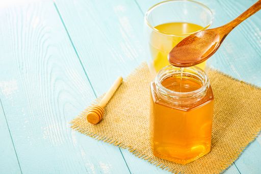 A closeup of golden honey pouring from a wooden spoon into a jar next to a juice glass