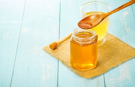 A closeup of golden honey pouring from a wooden spoon into a jar next to a juice glass