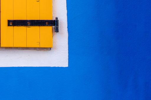 A close-up of a single window with a yellow shutter on a blue wall in the background