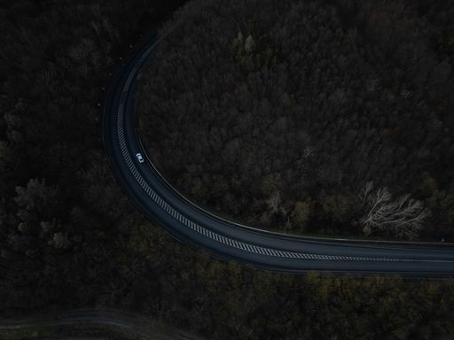 Bird's eye view of a wide road curving around a hill covered by dark trees in the wilderness
