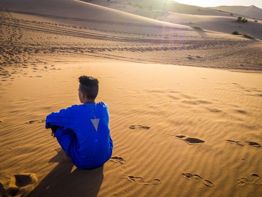 Bedouin boy wearing a blue djellaba and orange scarf standing in the desert
