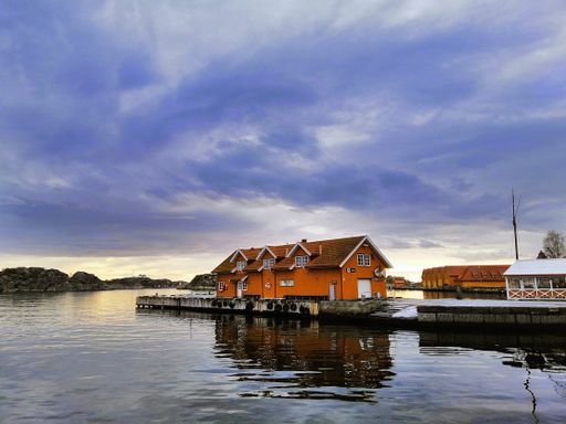 A red house surrounded by the sea and greenery under a cloudy sky during the sunset in Norway
