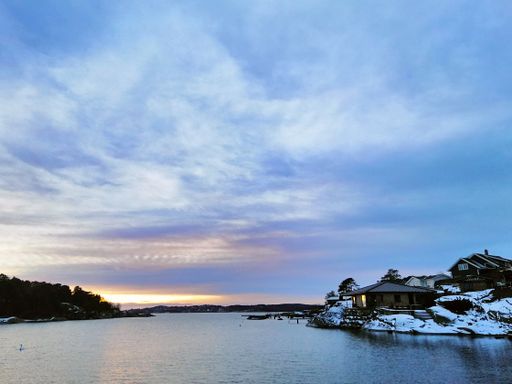 The sea surrounded by greenery and buildings covered in the snow during the sunset in Ostre Halsen