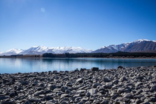 Lake Tekapo with blue sky and stones at the shore in foreground and snowy mountains in the back