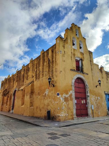 An old, yellow-painted building with a large red wooden door in Mexico with a cloudy sky in the background