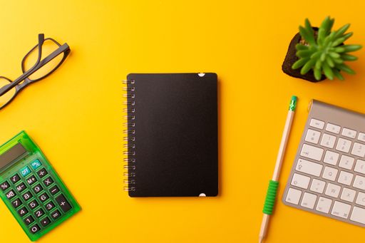 A top view of a journal, calculator, a pencil, keyboard, and a plant on a yellow background