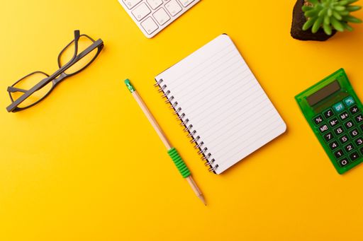 A top view of a journal, calculator, pencil, glasses, keyboard, and a plant on a yellow background