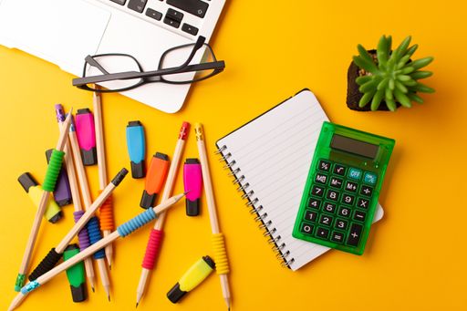 A top view of a laptop, journal, calculator, pencils, colorful highlighters, a plant, and glasses