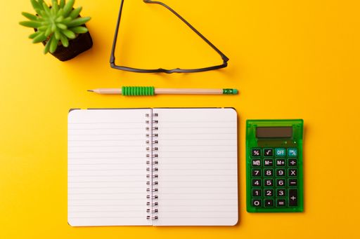 A top view of a journal, calculator, glasses, a pencil, and a plant on a yellow background