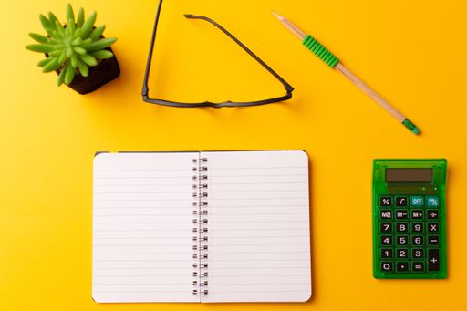 A top view of a journal, calculator, glasses, a pencil, and a plant on a yellow background
