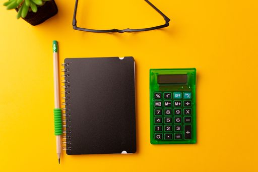 A top view of a journal, calculator, glasses, a pencil, and a plant on a yellow background
