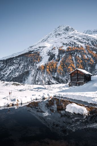 Swiss chalet in a wintry landscape with golden larches and snowy mountains reflecting in a small mountain lake in Arolla, Wallis, Switzerland