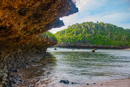 View on smooth wavy and reflecting sea water to a green hill and cloud sky, flanked from a brown fissured rock.