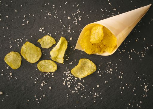 A top view of fried crunchy potato chips with salt spilling out of paper cone on a table surface