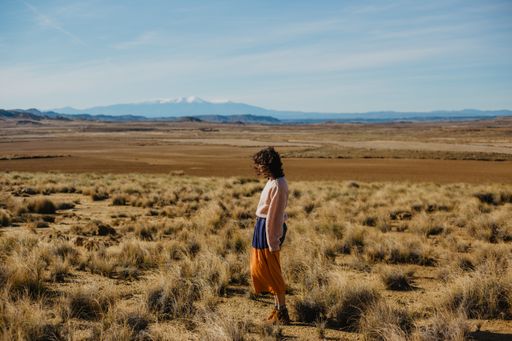 A woman in a long sleeve sweater and a long skirt standing a large brownfield with dried grass