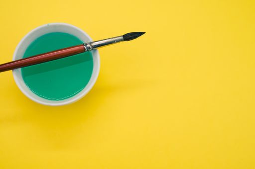 A white container with green liquid paint and a paintbrush on a yellow surface - copy space