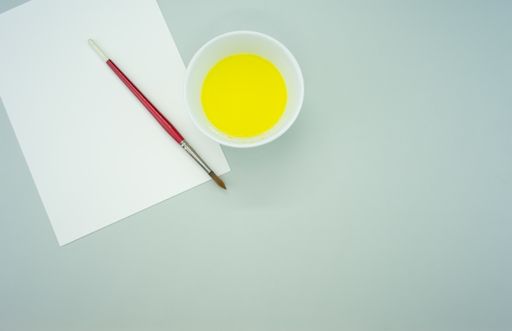 A white container with yellow paint, paintbrush and a blank paper on a green surface - copy space