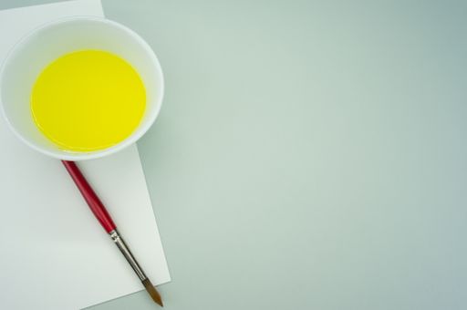 A white container with yellow paint, paintbrush and a blank paper on a green surface - copy space